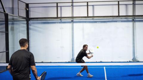 Two men playing padel. One of the men is smiling and laughing as he goes to hit the ball.  