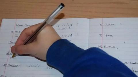 A closeup of a child's hand, as they write with a black biro on a school book, they wear a blue jumper.