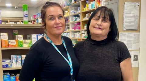 Two women stand in a pharmacy, one with a blue top and jeans, the other with a black dress