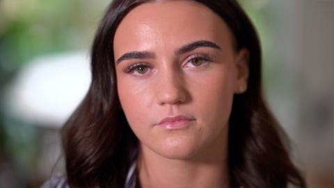 A young woman with long brown hair looks into the camera with a serious expression