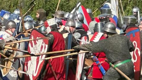A large number of men in medieval armour carrying spears and shields taking part in a mock battle with red shields