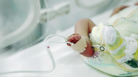 A stock image of a premature baby lying in an incubator in hospital, wearing a baby grow. It is a close up of the baby's legs with medical tubes coming from its foot.
