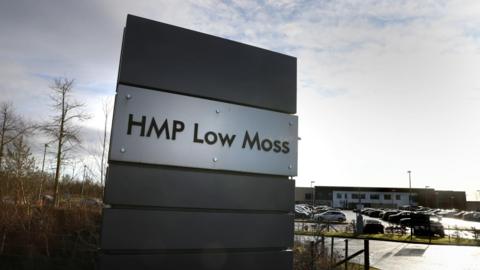 HMP Low Moss sign at its entrance with buildings and a car park full of cars in the background