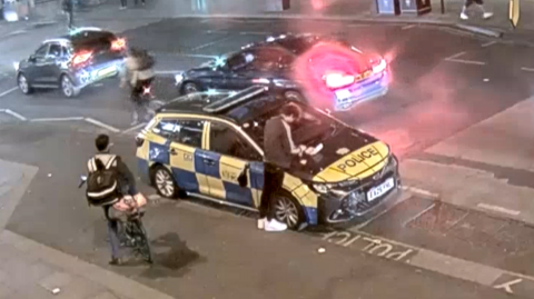 A man can be seen leaning over a police car with a takeaway tray placed on the bonnet. 