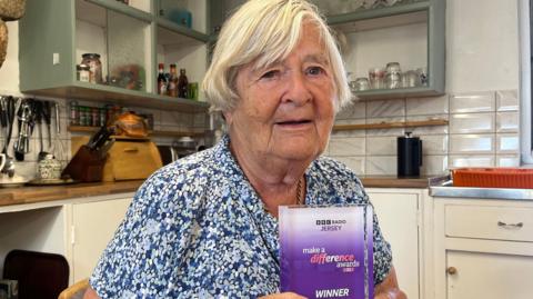 Genette sits at a wooden table in a homely kitchen, holding a purple “Make a Difference Awards – Winner” trophy. Behind her are open shelves, kitchen utensils, and a traditional stone feature above the cooker.