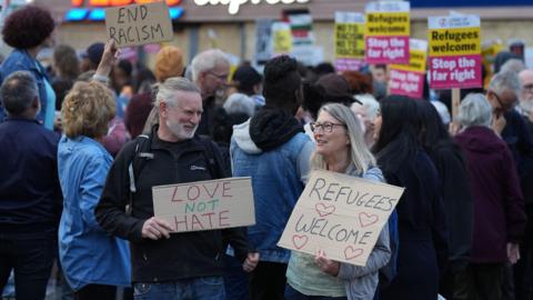 A man wearing a black coat holding a banner that says 'Love not hate'. A woman is standing next to him, wearing glasses, with a banner that says 'Refugees Welcome'. They are amongst crowds of people holding banners in an anti-racism protest