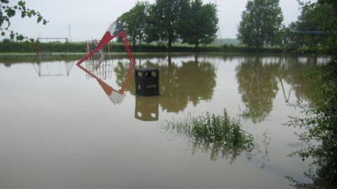 A flooded park with play equipment, including a slide and swings, in the distance. A bin is in the centre of the photo. Green trees and bushes surround the park. 