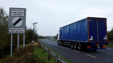 A blue truck drives away from the camera on the right of the image. It is on a quiet road. On the left of the image is a sign, which reads 'Welcome to NORTHERN IRELAND - Speed limits in miles per hour'. The sky is grey and overcast. 