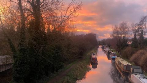 A canal runs through the picture with several barges moored up on the right-hand side. There is a footpath and a lot of trees on the left-hand side of the canal. Overhead, the sky is a golden yellow colour as the sun sets.