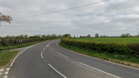 A curved section of rural road with the bend signage visible. There are hedgerows and fields on either side of the road. 
