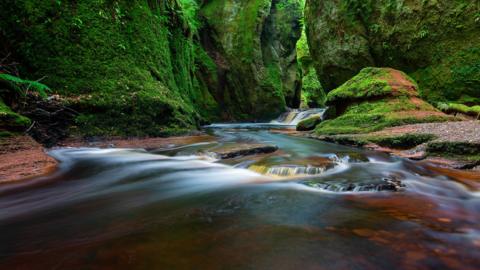 A long exposure shot of water flowing down a steep gorge. A large rock can be seen in the foreground and steep green covered rocks can be seen in the background 