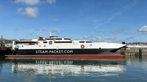 The Manannan ferry in Douglas Harbour. It is white, red and black and has STEAM-PACKET.COM on the side. The sky above is blue.