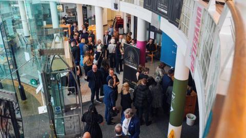 A crowd of people can be seen from above standing inside a building. There is a door to the left and columns through the middle. A large black sign is in front of the doors.