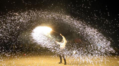 A man spins a fire stick around above his head, spraying sparks out around him