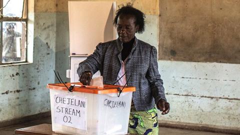 A woman in a patterned jacket and African wrap for a skirt stands at a ballot box putting her vote in. She is in an empty classroom and a blackboard can be seen behind her.