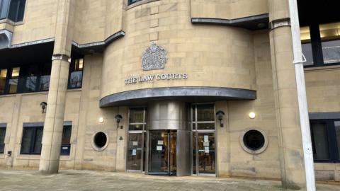 A stone building with a sign and emblem saying THE LAW COURTS above a steel revolving door. On either side of the door are two round windows and two large stone columns.