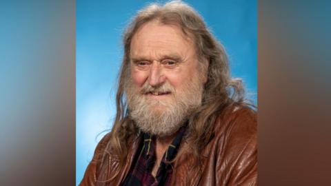 Wild Young, who ha long brown hair and a greying beard and is wearing a brown leather jacket over a dark checked shirt. The background it light blue.
