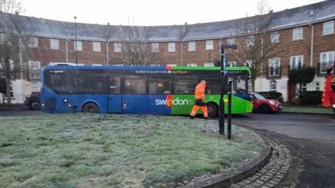 A view of a bus and a grass-covered roundabout. The bus is stopped and a man in an orange high-viz jacket is stood in front of the bus. Behind are parked cars and a modern row of houses.