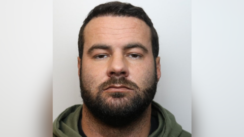 Mugshot of Oliver Cooper. He is pictured against a plain background with a blank expression. He has short dark hair and facial hair. 