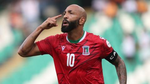 Emilio Nsue in red Equatorial Guinea shirt and with captain's armband points with his right finger at his temple.