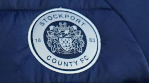 A silver and blue version of Stockport County's badge, which bears the club's name and the crest of two rampant lions either side of a shield with a crowned helmet above
