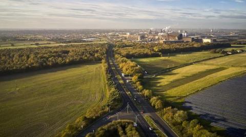 Aerial view of Mortal Ash Hill with trees along the side of the road and the town's steelworks in the distance.