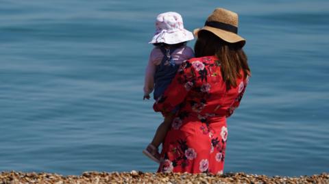 A woman is holding a young girl in her arms with their backs turned to the camera. They are on the beach looking at the clear blue water and there is shingle in the foreground. The woman is wearing a red floral dress and large floppy straw hat and the child is wearing a blue pinafore over a pink top and a white and pink patterned sun hat.