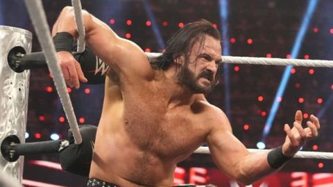 Drew McIntyre in the ring, crouching by the turnbuckles, with one arm resting on the ropes. His face is in a grimace, while he is wearing black trunks and boots. He appears about ready to pounce on an opponent.