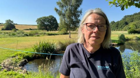 A woman with shoulder-length grey hair, wearing glasses and a dark blue t-shirt. She is stood looking into the camera. Behind her is a pond, grass and trees