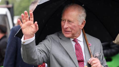 King Charles smiles and waves while sheltering under an umbrella 
