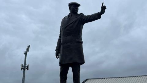 A statue of a man, wearing a flat cap and a jacket, standing sideways while holding out his arm with his index finger pointing up.