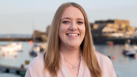 Poppy Murray with dark blonde hair and a pink shirt  smiling at the camera in front of Castle Cornet in St Peter Port