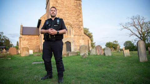 PC Dan Counsell wears black trousers and a black police vest with his walkie talkie attached to his chest. He has short dark hair and stubble. He is standing with his arms folded in front of a medieval church made of stone. There are grave stones around him. 