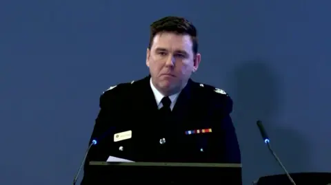 Man in black uniform, white shirt and black tie in front of blue background