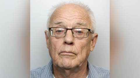 David Kuca has black-rimmed glasses. short grey hair, and is stood in front of a grey wall.