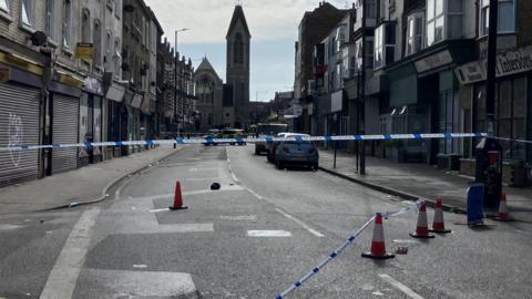 A shot of Northdown Road in Cliftonville closed off by blue and white police tape with police cars in the distance