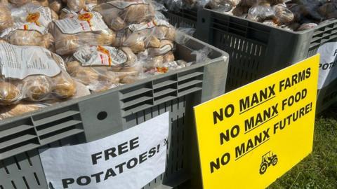 A large grey container filled with clear plastic bags of potatoes. A sign saying 'free potatoes' is attached to the box. A yellow placard says 'no Manx farms, no Manx food, no Manx future!'