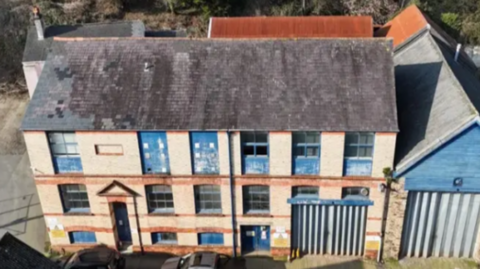 The picture shows a large, old industrial-style building photographed from above. The structure has three floors, with pale brick walls and bright blue window frames and doors. The roof is slate and shows patches of weathering. The ground floor has wide shutter-style doors on the right side that look suited to vehicles or workshop use. 