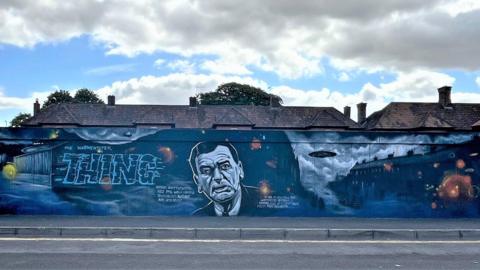 A wall painted with a mostly dark blue mural, with roofs of houses behind the wall. The mural reads 'The Warminster Thing' and includes a flying saucer near a row of terraced houses. A portrait of journalist and ufologist Arthur Shuttlewood is included prominently. 