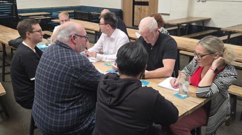 A group of people sit around a bench in a brewery, with several writing in notepads.