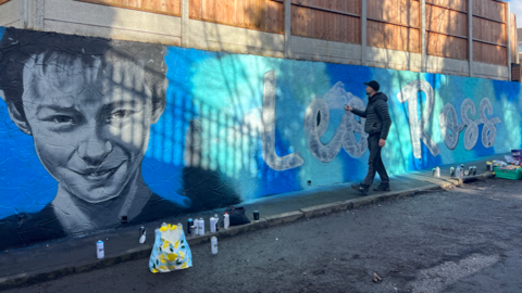 A man touches a wall on which a gigantic blue mural has been painted. On the left is Leo Ross's face, neck, and shoulders. On the right is his name, painted on the wall in white writing.