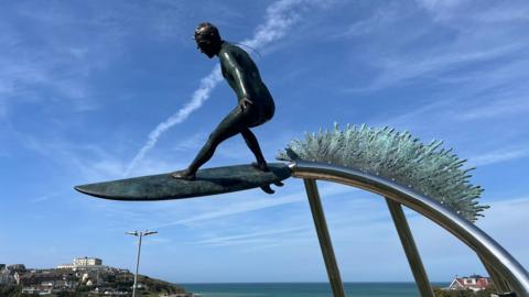 A statue of a surfer on a surf board suspended on a steel pole with the sea behind it. Behind the surfer is a bronze wave symbol