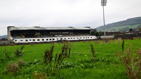 Overgrown grass in front of a stadium. Hills are in the background. 