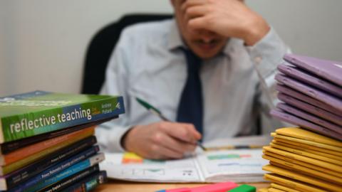 Teenage boy in shirt and school tie writing in a workbook at a desk with pens and other work books piled on it