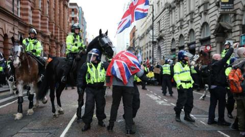 Police officers on horseback and with helmets. A man with a union jack flag wrapped around him is near to the officers. A crowd of people holding signs can be seen in the background.