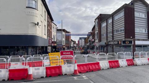 In the foreground is a row of red and white barriers blocking access to the road beyond it, which is flanked by buildings.