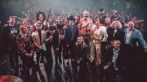 Twenty people - performers at a concert - smile at the camera. They are a mix of men and women, two people are holding microphones and one a guitar. A large crowd of fans can be seen behind them. 