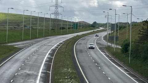 A general view of the A465 dual carriageway, which has two lanes travelling in each direction separated by a grass verge. Lampposts line each side of the road, with an electricity pylon on the left hand side and a hill beyond.  