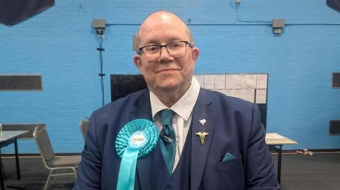A man in a navy blue suit and tie is smiling at the camera, he is wearing glasses and a bright blue rosette