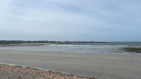 Two people walk on the beach at Vazon Bay on a cloudy day. Part of the sandy beach had water on it from a previous tide.